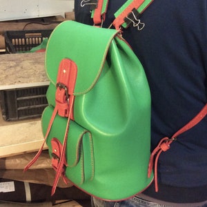 May include: A green leather backpack with red leather straps and buckles. The backpack has a small pocket on the front.