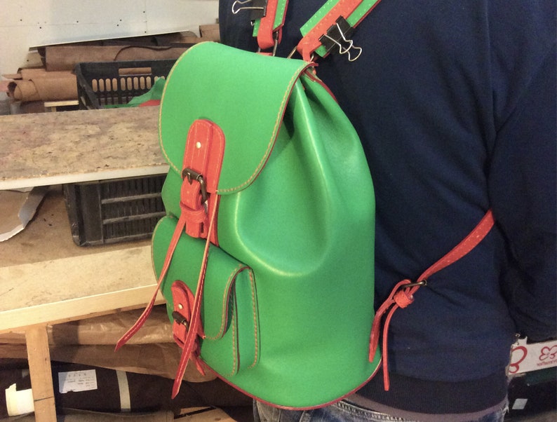 May include: A green leather backpack with red leather straps and buckles. The backpack has a small pocket on the front.