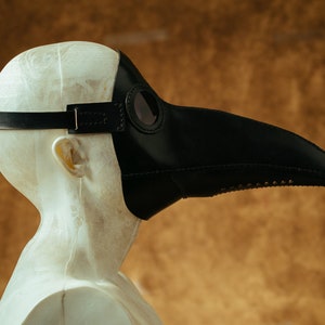 May include: A black leather plague doctor mask with a long beak and a round eye piece. The mask is worn on a mannequin head.