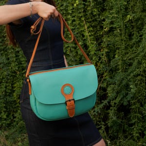 May include: A turquoise satchel bag with a brown leather strap and buckle. The bag has a rounded flap closure and a contrasting brown trim. The bag is held by a person in a denim dress against a backdrop of green foliage.