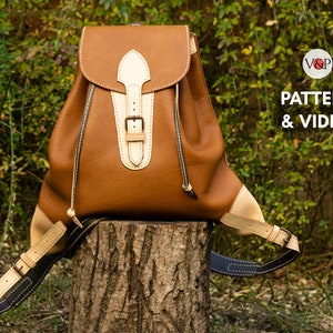 Puede incluir: Una mochila de cuero marrón con una solapa y una hebilla de cuero blanco. La mochila está sentada sobre un tocón de árbol. El texto "PATTERN & VIDEO" es visible en la esquina superior derecha de la imagen.