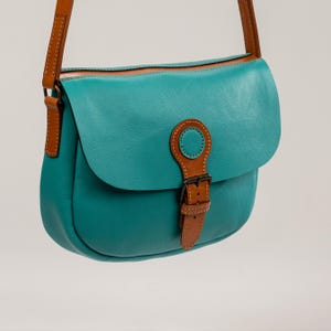 May include: A teal leather crossbody bag with a brown leather strap and accents. The bag features a flap closure with a buckle and a circular accent. The bag is designed with a saddlebag style.