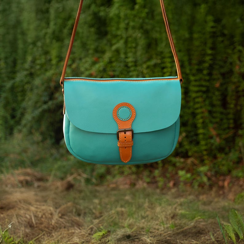May include: A turquoise leather crossbody bag with a brown leather strap and buckle. The bag has a rounded flap and a contrasting brown trim. The background is blurred, with green foliage and brown grass.