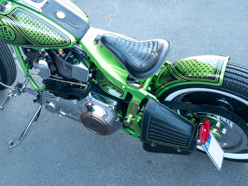 May include: A green motorcycle with a black leather seat and a black leather saddlebag. The motorcycle has a chrome engine and a white wall tire. The motorcycle has a green and black paint job with a fish scale pattern.