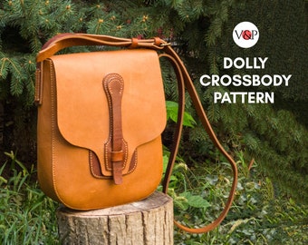 May include: A brown leather crossbody bag with a flap closure and a decorative strap. The bag is sitting on a tree stump in front of green foliage. The text "DOLLY CROSSBODY PATTERN" is visible in the image.