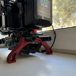 May include: A red camera support stand with two black handles. The stand is designed to support a camera and has a curved shape. The stand is made of plastic and has a smooth surface. The stand is sitting on a white surface. The camera is not visible in the image.