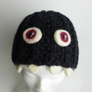 Könnte beinhalten: Schwarze Strickmütze mit großen weißen Filzaugen und roten Pupillen. Die Mütze hat eine Reihe weißer Filzzähne am unteren Rand.