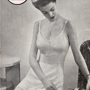 May include: A black and white photo of a woman wearing a white crocheted cami-knickers outfit. The outfit is made with a lacy stitch and has a fitted bodice and a short skirt. The woman is sitting on a chair with a floral arrangement in front of her. The image includes the text "Knitpat No. 26 CAMI-KNICKERS 3d."