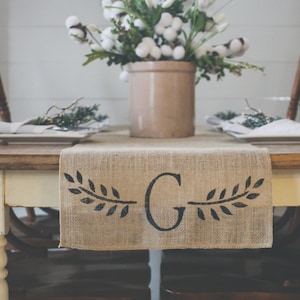 May include: A burlap table runner with a black letter "G" and two black leafy branches on either side of the letter. The runner is on a wooden table with two chairs on either side. A vase with white cotton flowers is in the center of the table.