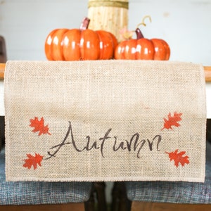 May include: A burlap table runner with the word "Autumn" written in black script and red leaf accents. Two orange pumpkins are in the background.