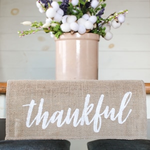 May include: A burlap table runner with the word "thankful" printed in white. The runner is on a table with two gray cushions in the background. A vase with white cotton and purple flowers is in the background.