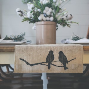 May include: A burlap table runner with a silhouette of two birds perched on a branch. The runner is brown and has a natural texture. The birds are black and are facing each other.