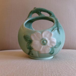 May include: A light green ceramic basket with a rounded body and a curved handle. The front features a raised, white five-petaled flower with green leaves. The design includes open spaces between the handle and the main body of the vessel.