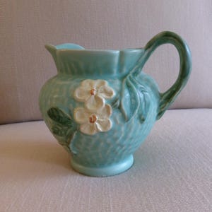 May include: A light blue ceramic pitcher featuring a textured surface with embossed white flowers and green leaves. The pitcher has a curved handle, a scalloped rim, and a rounded body that tapers toward a circular base, finished in a glossy glaze.