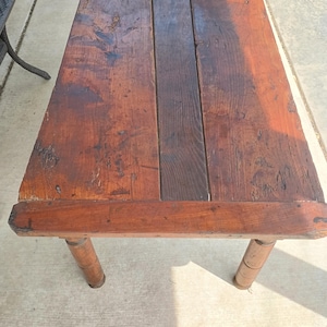 May include: A rectangular wooden table with a dark brown finish. The tabletop is constructed from several planks of wood, and the table has four round legs. The table appears to be antique or rustic in style.