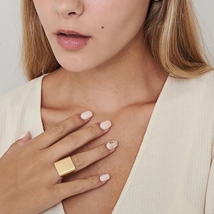 May include: A gold square ring worn on a finger with light pink nail polish.