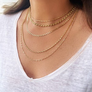 Könnte beinhalten: Eine mehrlagige goldene Kettenkette mit fünf verschiedenen Kettenstilen. Die Ketten sind alle golden und übereinander gelegt. Die Kette wird von einer Person mit einem weißen Hemd getragen.