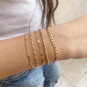 May include: Five gold chain bracelets stacked on a person's wrist. The bracelets are of different widths and styles.