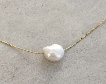 May include: A delicate gold-tone chain necklace featuring a single, irregular, off-white baroque pearl centered on the strand. The chain has a fine, textured link design, and the pearl displays a smooth, lustrous surface with a unique organic shape.