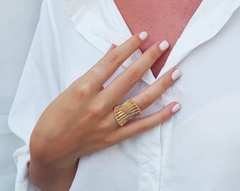 May include: A gold ring with a geometric design. The ring is worn on the index finger of a hand.