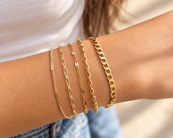 May include: A set of five gold-toned metal bracelets worn together. The collection includes a thin curb chain, an infinity link chain, a delicate oval link chain, a rectangular bar link chain, and a thicker, classic curb chain link bracelet.