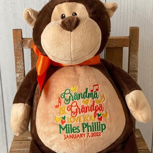 May include: A brown plush monkey with a tan face, ears, hands, and feet, wearing an orange ribbon. The monkey's belly features embroidered text: "Grandma & Grandpa LOVE YOU Miles Phillip JANUARY 7, 2022". The monkey is sitting on a wooden chair.