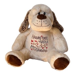 May include: A plush dog toy with cream-colored fur, brown ears, and brown paw pads. The chest features embroidered text that reads Heaven sent with a kiss from my Grandma, accented with small red hearts in brown and red thread colors.