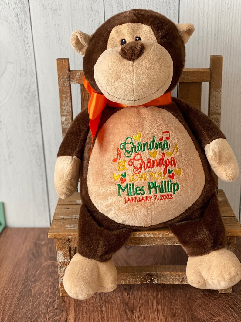 May include: A brown plush monkey with a tan face, ears, hands, and feet, wearing an orange ribbon. The monkey's belly features embroidered text: "Grandma & Grandpa LOVE YOU Miles Phillip JANUARY 7, 2022". The monkey is sitting on a wooden chair.