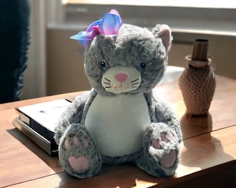 May include: A plush gray cat stuffed animal with a soft, fuzzy texture, featuring a white belly patch, a pink embroidered nose and mouth, and pink paw pads. A large, multi-colored blue and pink hair bow is attached to the top of its head.