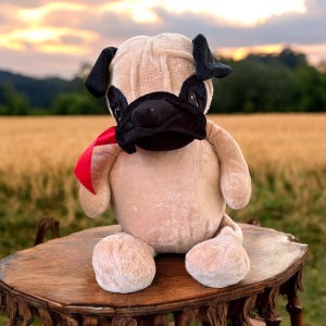 May include: A plush pug toy with light tan fur, a black muzzle, and black ears. The stuffed animal features a bright red ribbon tied around its neck, with a soft, rounded body and limbs, designed to sit upright on a flat surface.