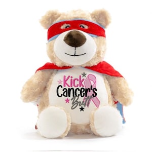 May include: A plush teddy bear wearing a red superhero mask and cape. The bear has a brown nose and is embroidered with the text "Kick Cancer's Butt" and a pink ribbon.