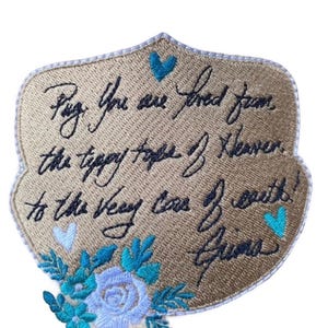 May include: A shield-shaped embroidered patch with a gold background and a white border. It features black cursive text reading: "Pug, You are loved from the tippy top of heaven to the very core of earth! Grima". Includes blue and light blue hearts and flowers.