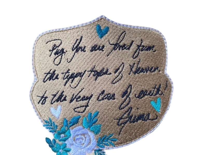 May include: A shield-shaped embroidered patch with a gold background and a white border. It features black cursive text reading: "Pug, You are loved from the tippy top of heaven to the very core of earth! Grima". Includes blue and light blue hearts and flowers.