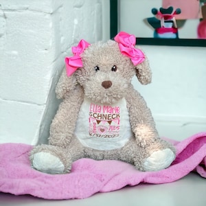 May include: A beige plush dog toy with two bright pink bows on its ears. The dog wears a white shirt embroidered with the name Ella Marie Schneck, birth details including 20 inches and 7lbs, and the date 8/17/2023 in pink and brown thread.