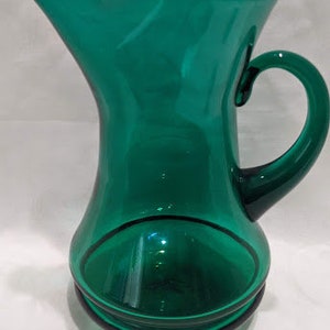 May include: A green glass pitcher with a wide mouth and a handle. The pitcher has a rounded body and a flared base.