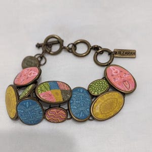 May include: A colorful bracelet with oval-shaped, patterned beads in shades of pink, yellow, blue, and green. The bracelet has a bronze-colored chain and clasp, with the brand name "JILZARAH" on a small tag.