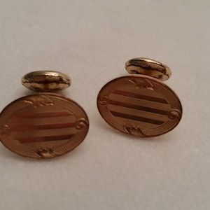May include: A pair of oval-shaped gold-toned cufflinks featuring a horizontal striped pattern across the center. Each cufflink has a decorative border with small engraved details and a rounded toggle backing for secure fastening to a shirt cuff.