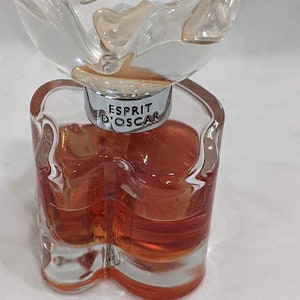 May include: A clear glass bottle of perfume with a flower-shaped top. The bottle is filled with a reddish-orange liquid. The label on the bottle reads "Esprit d'Oscar".