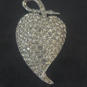 May include: A silver-toned brooch in the shape of a heart, encrusted with numerous clear, sparkling rhinestones. The brooch has a loop at the top, suggesting it could also be worn as a pendant. The heart shape is slightly asymmetrical.