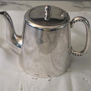 May include: A polished silver-toned teapot featuring a curved spout, a circular lid with a small spherical knob, and a C-shaped handle with decorative perforations at the top and bottom attachment points. The body has a smooth, reflective surface.