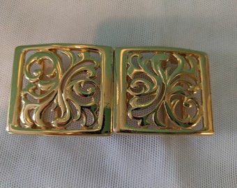 May include: A gold-toned metal belt buckle with a decorative, ornate, openwork design. The buckle is made of two pieces that connect together.