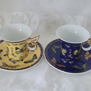 May include: Two decorative teacups and saucers. One cup and saucer is yellow with butterfly designs, the other is blue with butterfly designs. Both cups have gold-colored handles shaped like wings.