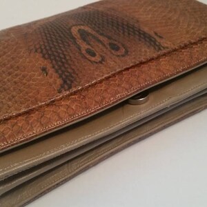 K&ouml;nnte beinhalten: Eine braune Leder-Clutch mit Schlangenhautmuster und einem silbernen Verschluss. Die Clutch ist in der Mitte gefaltet und hat ein beigefarbenes Futter.
