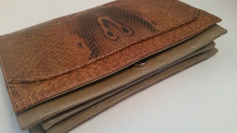 K&ouml;nnte beinhalten: Eine braune Leder-Clutch mit Schlangenhautmuster und einem silbernen Verschluss. Die Clutch ist in der Mitte gefaltet und hat ein beigefarbenes Futter.
