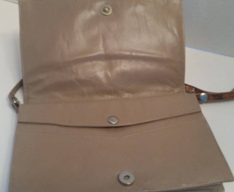 K&ouml;nnte beinhalten: Eine beige Lederhandtasche mit Klappenverschluss und einer kleinen Tasche auf der Vorderseite. Die Tasche hat einen braunen Lederriemen.