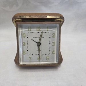 May include: A vintage Phinney-Walker alarm clock with a square face. The clock has a gold-toned metal casing and a light-colored face with black numerals and hands. The clock face reads "PHINNEY-WALKER" and "ALARM".