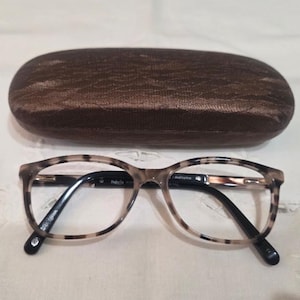 May include: Oval-shaped eyeglasses with a beige and black tortoiseshell pattern on the frames and black temples. The glasses are positioned in front of a dark brown, textured, oval-shaped hard-shell protective case with a subtle wavy pattern.