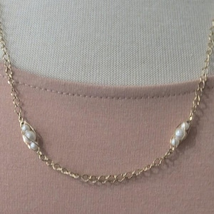 May include: A delicate gold-toned necklace featuring three small, white pearls encased in a woven gold setting. The necklace has a delicate chain and is displayed on a light pink background, suggesting a jewelry or accessory theme.
