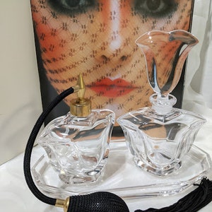 May include: Two clear glass perfume bottles with a floral design, one with a black and white tassel, on a clear glass tray. The bottles are shaped like a flower.