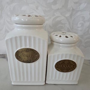 May include: Two white ceramic canisters with lids. The larger canister has a label that reads "COFFEE" and the smaller one reads "SUGAR". Both have a ribbed design and decorative oval labels.